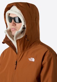 Chaqueta marrón con capucha y forro esponjoso, cierre de cremallera, cordones ajustables y un logo en el pecho. Presenta gafas de sol oscuras.