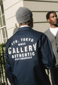 Námořnicky modrá bunda s bílým nápisem na zádech "GALLERY AUTHENTIC CRAFTSMANSHIP" a "LTD. TOKYO MMXX," doplněná texturovanou šedou čepicí.