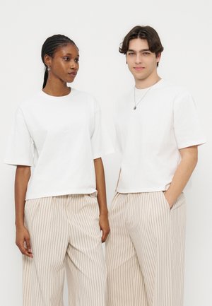 Δύο άτομα φορούν λευκές φαρδιές μπλούζες και μπεζ παντελόνια φαρδιές με ρίγες, στέκονται μπροστά σε μια απλή λευκή επιφάνεια.