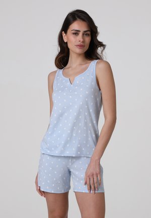 Donna che indossa un top smanicato azzurro chiaro e pantaloncini con piccoli motivi floreali bianchi, in piedi davanti a uno sfondo semplice.