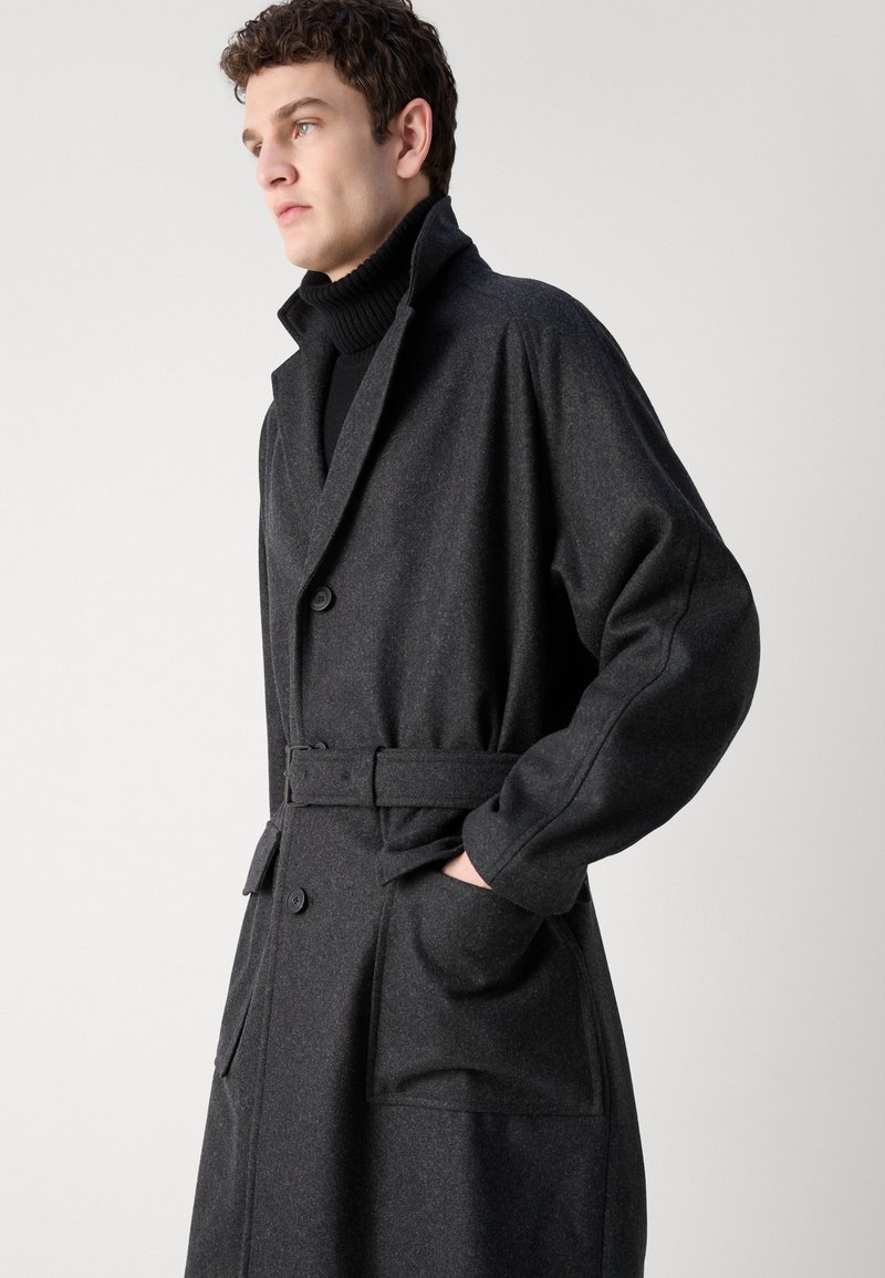 Manteau en laine gris foncé avec un design à double boutonnage, col surdimensionné, taille ceinturée et poches latérales. Tissu texturé avec une coupe décontractée.