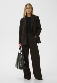 Donna che indossa un completo a gamba larga marrone scuro con una maglietta nera, mentre tiene una grande borsa tote nera, in piedi contro uno sfondo chiaro e uniforme.