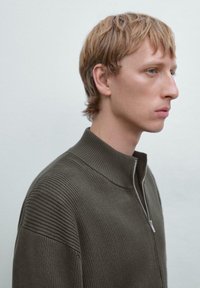 Giovane uomo con capelli biondi corti che indossa un maglione zip-up a coste verde scuro, di profilo a destra contro uno sfondo chiaro e uniforme.