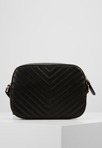 Lille sort kviltet læder crossbody taske med chevron-syninger, rektangulær form og sidelukning på hvidt displaypedestal.