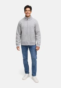 Grå strikjakke med lynlås og høj krave, sidelommer og ribbede manchetter, parret med blå jeans og hvide sneakers.