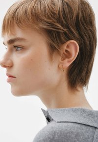 Aro de oro llevado en la oreja izquierda, cabello corto castaño, piel clara, vistiendo una camiseta gris con cuello y detalle de botones.