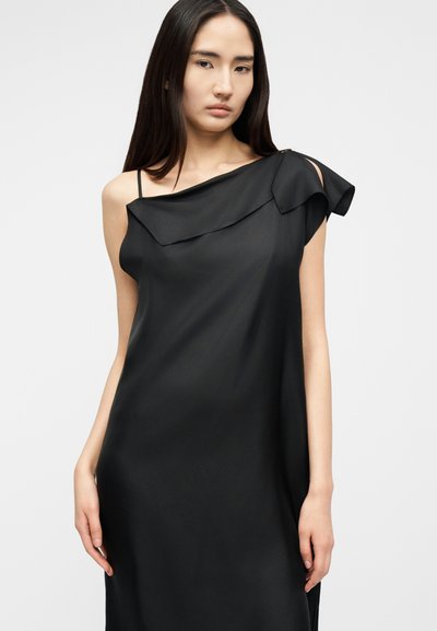 Femme portant une robe noire sans manches avec un décolleté asymétrique et un détail volanté sur une épaule, debout devant un fond uni.