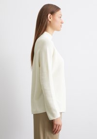 Crème-kleurige gebreide trui met een hoge halslijn, lange mouwen en een ontspannen fit, gepresenteerd tegen een neutrale achtergrond.