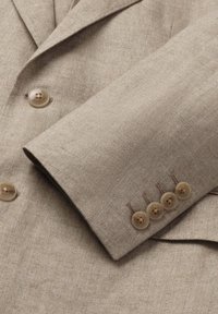 Detailný záber na rukáv béžového ľanového saka so štyrmi gombíkmi a predný panel s dvoma gombíkmi, zobrazujúci textúrovanú tkaninu a detaily šitia.