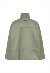 Lys olivengrøn langærmet jakke med høj krave og sort rektangulært mærke nær toppen i midten på ryggen.