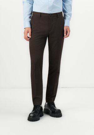 Hombre vestido con pantalones ajustados marrón oscuro, camisa de botones azul claro y zapatos negros gruesos de cordones, de pie frente a un fondo blanco.