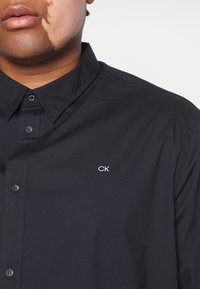 Personne avec une tache de naissance au visage portant une chemise noire boutonnée avec un petit logo blanc "CK" sur la poitrine.