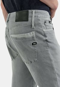 Jeans de mezclilla gris con un parche de cuero negro que presenta el nombre de la marca "GAS." Incluye dos bolsillos traseros y detalles de costura sutiles.
