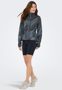 Giacca leggera di colore grigio scuro con texture a righe, zip frontale e vita elastica, abbinata a pantaloncini da bici neri e scarpe da ginnastica grigie.