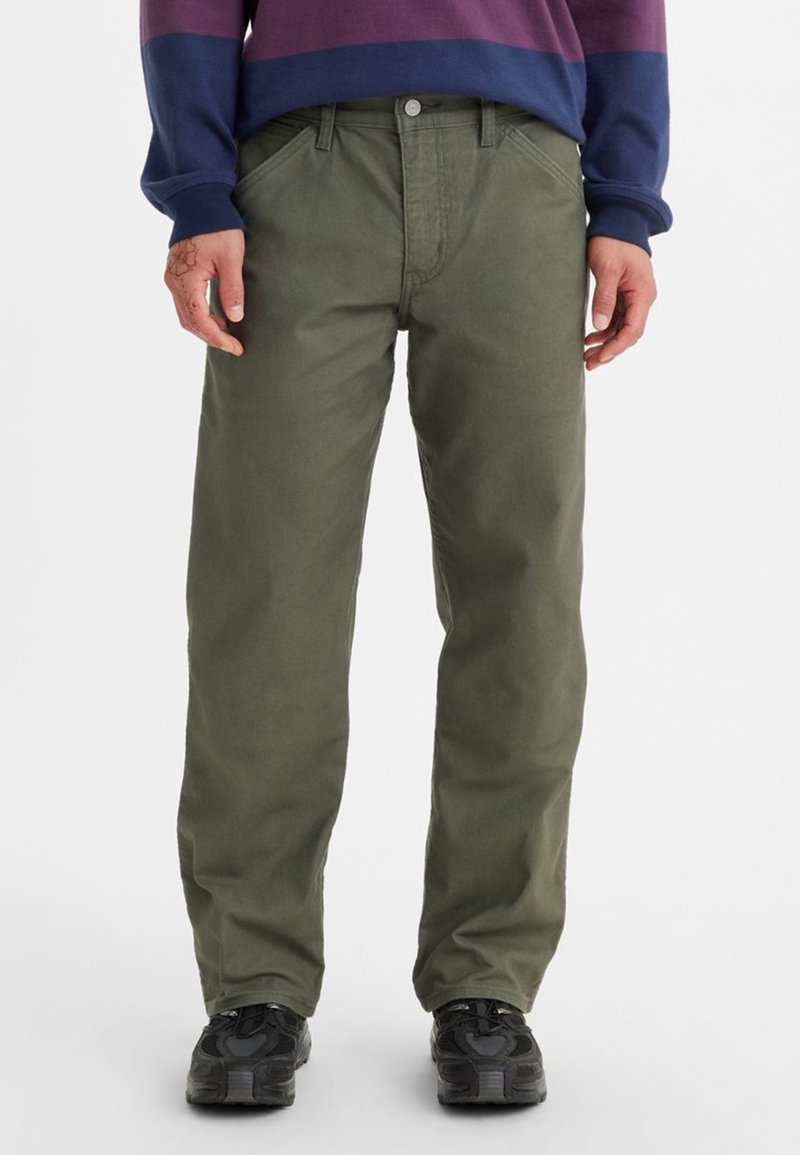 Homme portant un pantalon droit vert olive avec des poches avant et des baskets noires, debout devant un fond blanc.