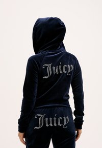Tuta in velluto blu navy con cappuccio, con "Juicy" in strass sul retro della giacca e dei pantaloni. Tessuto morbido e design aderente.