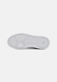 Fehér gumisbőr sneaker talp, amely texturált halszálkás mintával és kör alakú díszítésekkel rendelkezik. A "LACOSTE" márkaneve a sarkához közel nyomtatva.