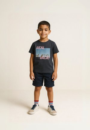 T-shirt di cotone grigio scuro con testo rosa "REAL VACAY" e grafica di una palma, abbinato a pantaloni corti blu navy e sneakers beige con dettagli blu.