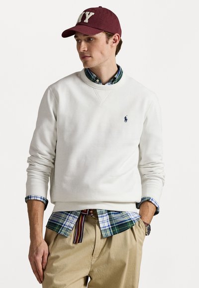 Jonge man die een witte sweater met ronde hals draagt over een groenblauw geruit overhemd, beige broek, bordeauxrode NY-pet en gestreepte riem, staand met één hand in de zak.