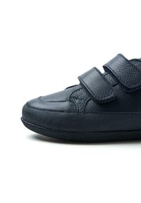 Scarpa nera in pelle con due cinturini in velcro, superficie testurizzata, punta arrotondata e suola in gomma. Il profilo laterale evidenzia le cuciture e i dettagli del design.