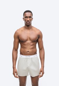 Muskularny mężczyzna stojący frontalnie, ubrany w jasnoszare bokserki Calvin Klein, na jednolitym białym tle.