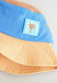 Bucket-Hut mit blauen oberen und orangefarbenen unteren Abschnitt, hergestellt aus strukturiertem Stoff, mit einem Gummi-Patch "RAD VIBES", das eine Palme zeigt.
