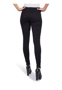 Mujer de pie con las manos en las caderas, vistiendo jeans ajustados negros y zapatos de tacón alto negros, de espaldas sobre un fondo blanco.