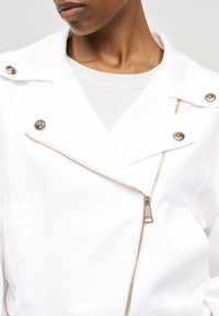 Κοντινό πλάνο ενός ατόμου που φοράει λευκό μπουφάν με χρυστό φερμουάρ και διακοσμητικά χρυσά κουμπιά, συνδυασμένο με λευκό εσωτερικό πουκάμισο.