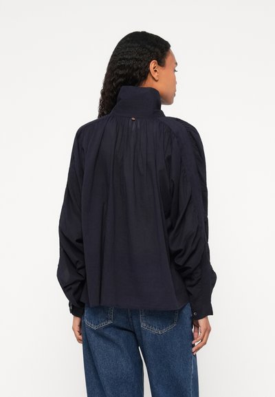 Maglietta a maniche lunghe blu navy con colletto alto, schiena arricciata e maniche oversize. Presenta polsini con bottone e una leggera lucentezza nella texture del tessuto.