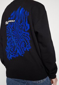 Sudadera negra con un patrón abstracto azul en la parte posterior y el texto "KARL LAGERFELD JEANS". Fabricada en material suave y con mangas largas.