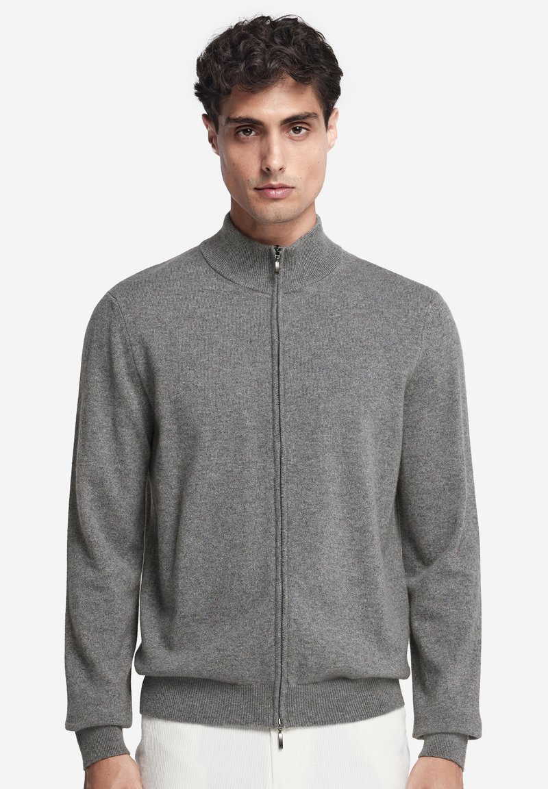 Mann trägt einen grauen Reißverschluss-Pullover mit hohem Kragen und gerippten Bündchen, steht vor einem schlichten weißen Hintergrund.