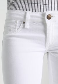 Jeans en denim blanc avec une coupe ajustée, dotés d'une fermeture à bouton, de poches traditionnelles et de détails de couture subtils. La texture est lisse et épurée.