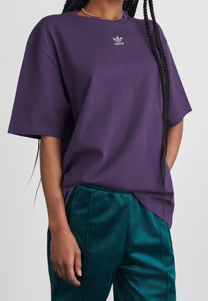Person iført en lilla Adidas t-shirt med korte ærmer og turkis velurbukser, stående mod en ensfarvet baggrund.