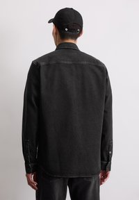 Man met een zwart denim shirt en een zwarte pet, staande met zijn gezicht naar een witte muur en zijn handen ontspannen naast zijn lichaam.