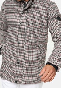 Tikkitakki houndstooth-kuosilla, jossa on punaisia yksityiskohtia, mustat napit ja taskut. Materiaali vaikuttaa pehmeältä ja lämpimältä.