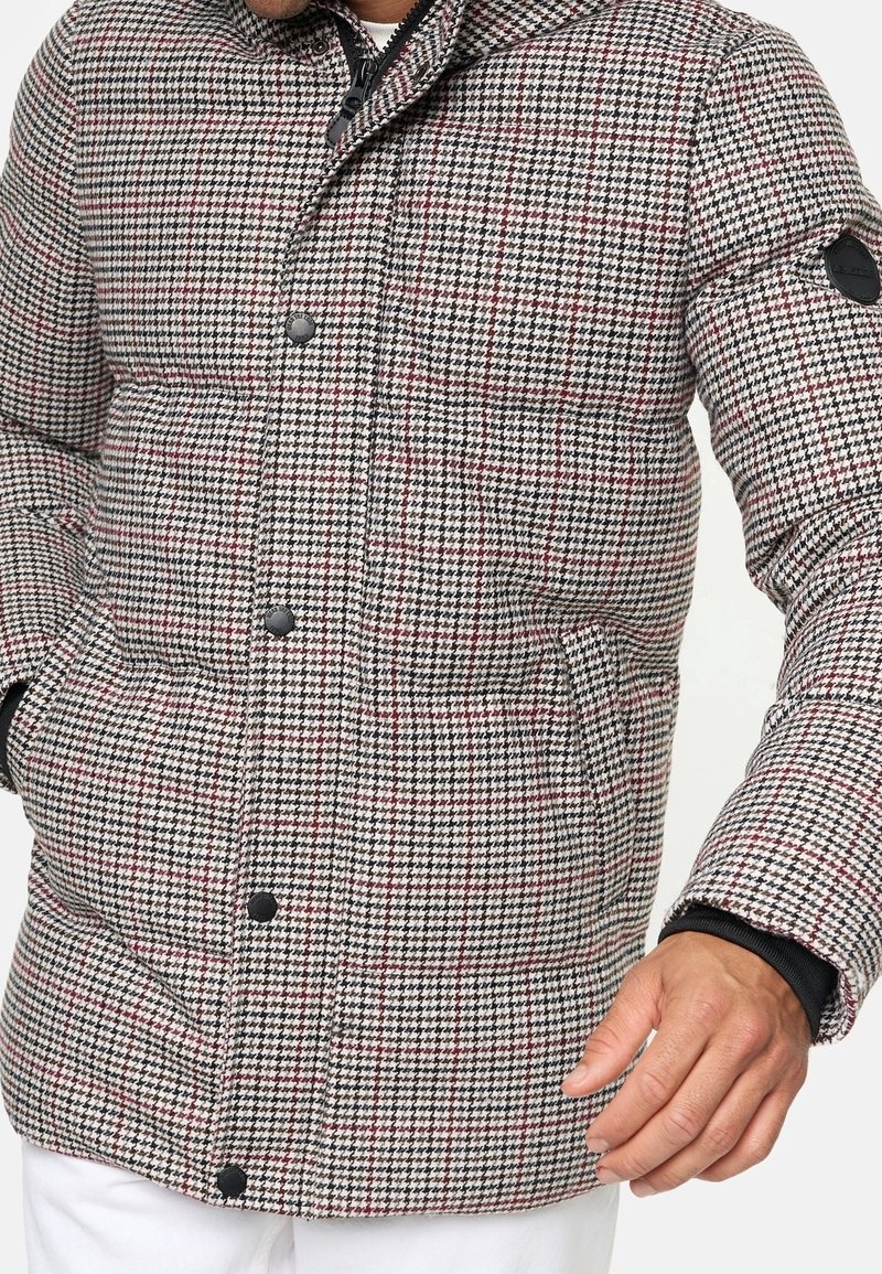 Tikkitakki houndstooth-kuosilla, jossa on punaisia yksityiskohtia, mustat napit ja taskut. Materiaali vaikuttaa pehmeältä ja lämpimältä.