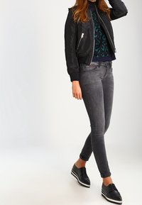 Giubbotto bomber nero sopra un maglione a righe verdi e blu, abbinato a jeans skinny grigi e scarpe nere con suole bianche.