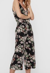 Tuta floreale con sfondo nero, caratterizzata da motivi di foglie rosa e verdi, spalline sottili e una vestibilità rilassata con gambe larghe.