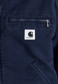 Close-up of navy blue Carhartt jacket pocket with horizontal zipper and white brand logo patch beneath it.