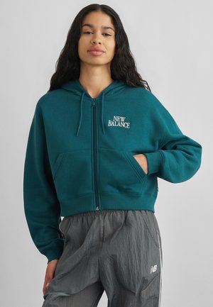 Nő, aki teal színű New Balance cipzáras kapucnis pulóvert és szürke New Balance melegítőnadrágot visel, egyik kezét zsebre téve áll, egyszínű háttér előtt.