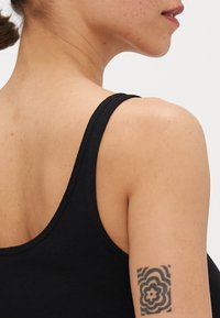 Svart tank topp med tynne stropper, som viser en jevn tekstur og en tettsittende passform. En blomstertatovering er synlig på overarmen.