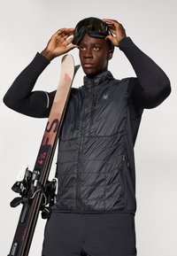 Melns pūkains vestes bez piedurknēm ar pilnu jostu, teksturēta priekšpuse un zirnekļa logo. Melna garām piedurknēm bāzes kārta un slēpes ar stiprinājumiem vienā rokā.