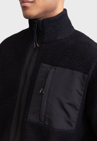 Jachetă fleece neagră cu guler înalt, buzunar din panou de țesătură netedă și texturi contrastante. Dispune de un fermoar frontal și hardware ajustabil.