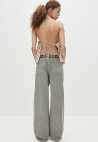 Una donna con i capelli ricci indossa un top halter senza schiena e jeans grigi sbiaditi a gamba larga con una cintura nera a occhielli, in piedi davanti a uno sfondo semplice.