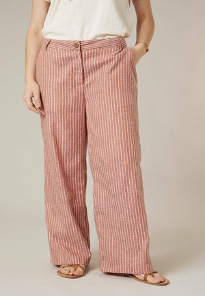 Pantalon à jambes larges avec des rayures blanches verticales sur un tissu rouge sourd, porté avec un haut blanc et des sandales couleur noisette.