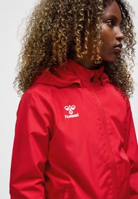Red waterproof jacket with a hood, featuring a zip closure, textured accents, and a white logo. The fabric appears smooth and lightweight.
