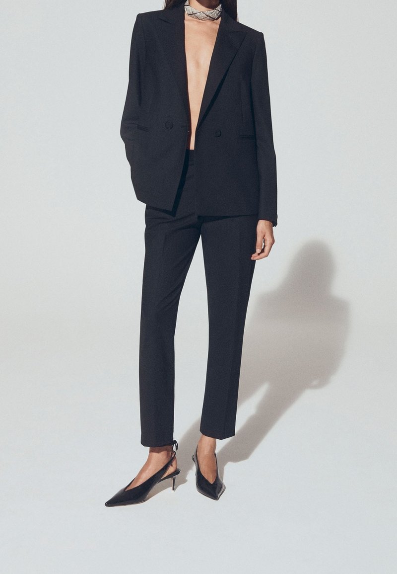 Blazer noir sur mesure et pantalon assorti en tissu élégant. Associés à des escarpins noirs pointus et un foulard de cou décoratif.