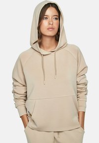 Vrouw met lichte huid en bruin haar draagt een beige hoodie met haar handen in de zakken, kijkt iets naar rechts, neutrale achtergrond.