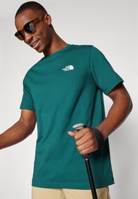 Tricou din bumbac de culoare teal cu un mic logo alb The North Face, purtat cu un baston de trekking în mâna dreaptă și ochelari de soare.
