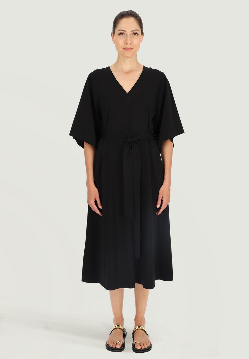 Czarna midi sukienka z dekoltem w serek, luźnymi rękawami oraz paskiem wiązanym w talii. Modelka zestawia ją z płaskimi sandałami z złotymi akcentami.